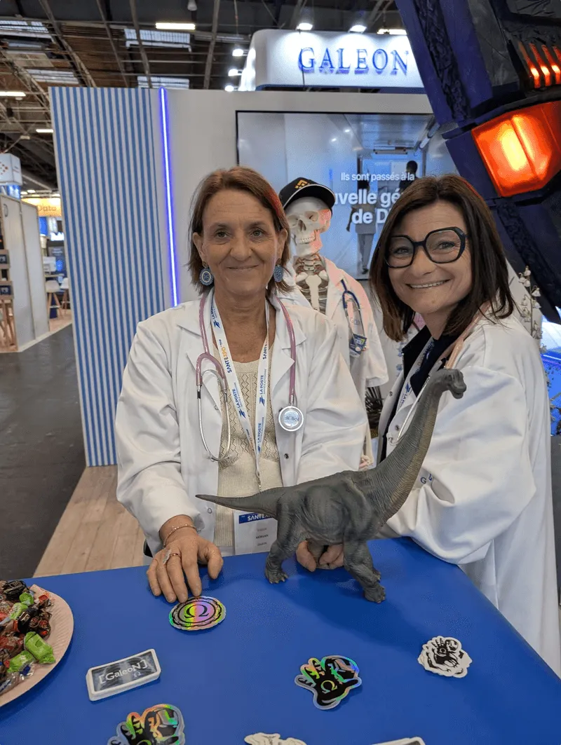 Two women in white coats with stethoscopes at the Galeon booth at SantExpo, with a toy dinosaur