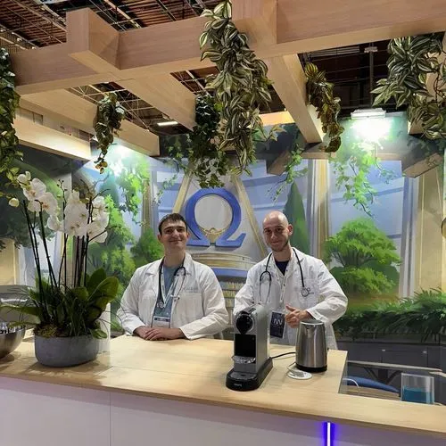 Two Galeon team members in white coats at an exhibition booth decorated with plants and the Omega logo